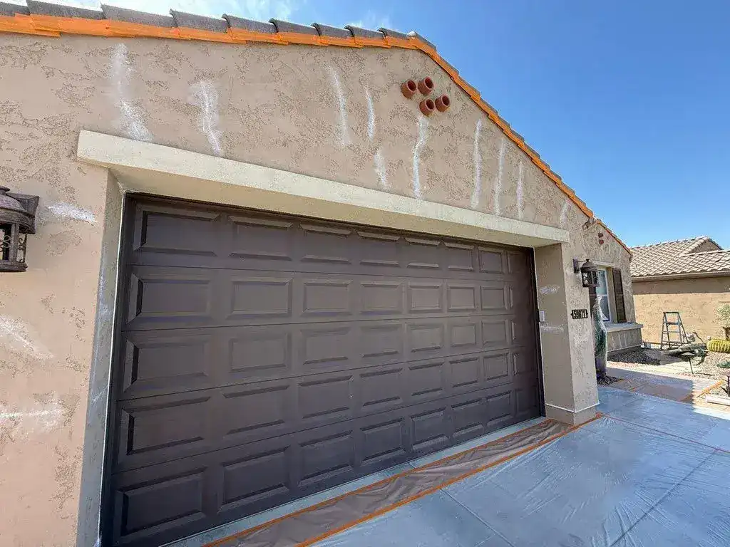 Stucco wall above a brown garage door with white patch marks where cracks were sealed, the driveway covered for painting