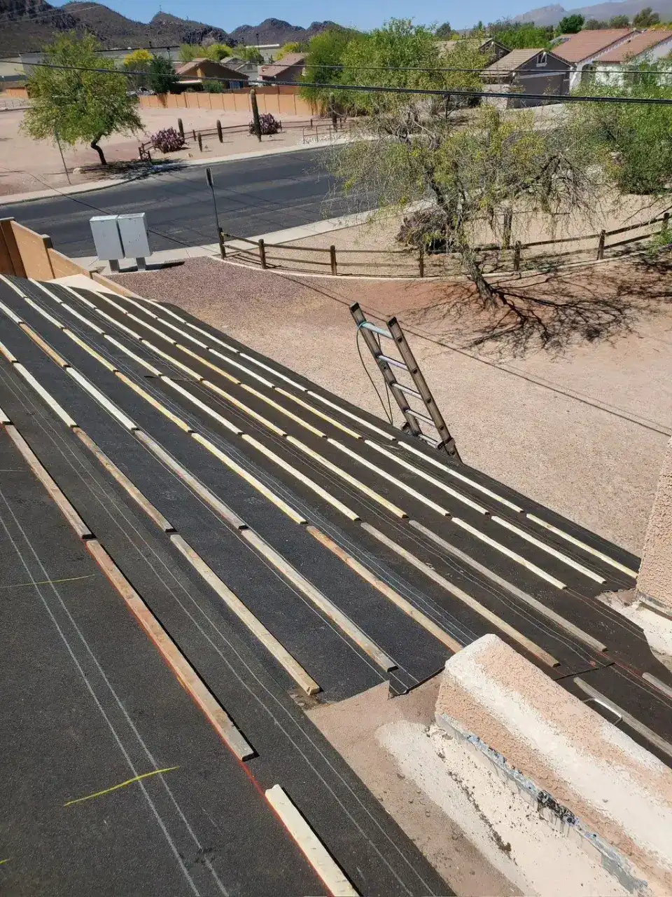 Roof deck covered in dark underlayment with rows of wood battens, a ladder at the edge and desert mountains in the distance